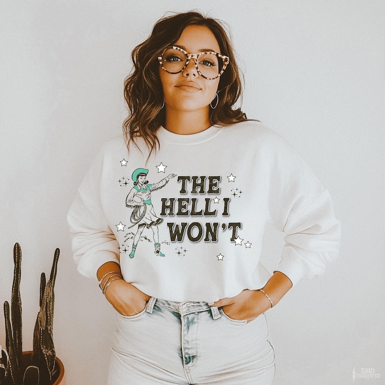 Puede incluir: Sudadera color crema con un gr&aacute;fico de vaquera y el texto "THE HELL I WON'T" en negro. La vaquera lleva un sombrero y botas turquesas, sosteniendo un lazo. Estrellas rodean el texto y el gr&aacute;fico.