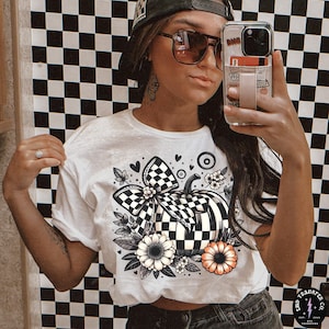 May include: White t-shirt with a black and white chequered pumpkin design, bow, flowers, and hearts. The pumpkin is the focus, with a black stem and decorative bow. The shirt has a crew neck and short sleeves.