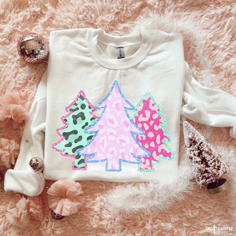 May include: A white sweatshirt featuring three Christmas trees with leopard print patterns in teal, pink, and fuchsia. The trees are outlined in glitter. The sweatshirt is on a pink, fluffy surface.