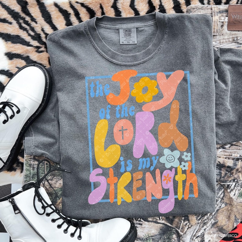 May include: A gray long-sleeve shirt with the text "the Joy of the Lord is my Strength" in colorful, retro-style lettering. The shirt is displayed with white boots and camouflage pants.