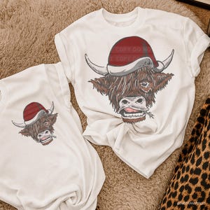 May include: Two white t-shirts featuring a Highland cow wearing a Santa hat. The cow has brown fur, white horns, and a sprig of grass in its mouth. The Santa hat is red with a white trim. The shirts are on a brown carpet.