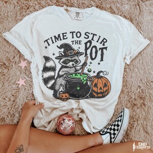 May include: Cream-colored t-shirt with a Halloween-themed graphic. The design features a raccoon in a witch hat stirring a cauldron, with the text "TIME TO STIR THE POT". A jack-o'-lantern and stars complete the design.