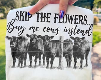 Könnte beinhalten: Weißes T-Shirt mit dem Text "SKIP THE FLOWERS... Buy me cows instead" in schwarzer Schrift. Darunter eine Schwarz-Weiß-Illustration von fünf Kühen hinter einem Zaun.