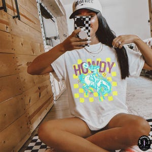 May include: White t-shirt with a graphic of a cowboy riding a horse. The graphic is in blue, yellow, and green. The text "HOWDY" is printed in maroon above the graphic.