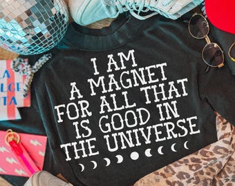 May include: A black long-sleeve shirt featuring the white text I AM A MAGNET FOR ALL THAT IS GOOD IN THE UNIVERSE with a moon phase graphic below. The shirt is surrounded by a disco ball, light blue sneakers, sunglasses, and a leopard print scarf.