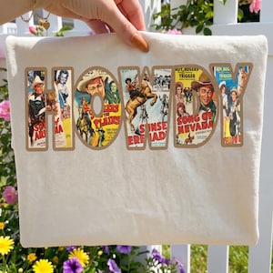 May include: A white t-shirt featuring the word HOWDY in large block letters. Each letter contains a collage of vintage Western movie poster imagery, including cowboys, horses, and titles like Song of Nevada and Riders of the Plains.