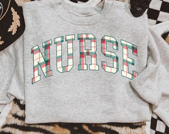 以下が含まれることがあります： 「NURSE」の文字が大きくチェック柄でプリントされた、ヘザーグレーのスウェットシャツ。チェック柄は赤、緑、クリーム色を使用。スウェットシャツは模様のある表面に表示されています。
