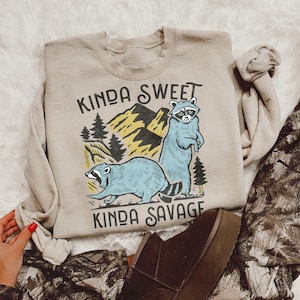 May include: A tan sweatshirt with a graphic of two raccoons in a forest setting. The text "Kinda Sweet Kinda Savage" is printed on the sweatshirt.
