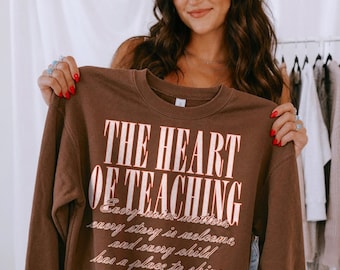 May include: A dark brown crewneck sweatshirt featuring the text THE HEART OF TEACHING in large, light-colored capital letters. Below, smaller cursive text reads Every voice matters, every story is welcome, and every child has a place to shine.