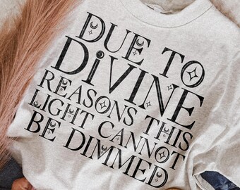 May include: A light gray crewneck t-shirt featuring black text that reads DUE TO DIVINE REASONS THIS LIGHT CANNOT BE DIMMED, accented with small star and crescent moon symbols interspersed throughout the lettering in a bold, serif typeface.