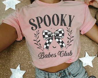 Może przedstawiać: Jasnoróżowa koszulka z napisem "SPOOKY Babes Club" w kolorze czarnym. Projekt zawiera szachownicową kokardę, gałązki i małego nietoperza. Koszulka wykonana jest z miękkiego materiału.
