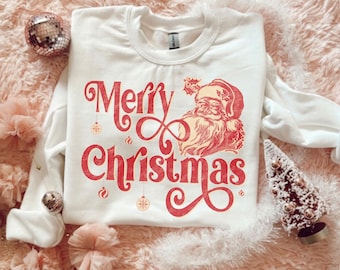 May include: A white crew neck sweatshirt featuring a red "Merry Christmas" design with a Santa Claus illustration. The festive graphic includes hanging ornaments and decorative flourishes, ideal for the Christmas holiday.