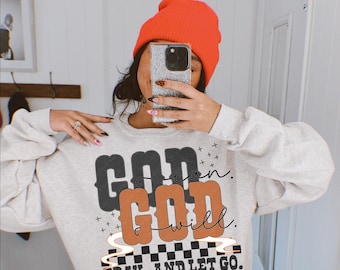 May include: A light gray sweatshirt with the text "GOD can, GOD will, PRAY, AND LET GO." The text is in black and orange, with a checkered pattern at the bottom. The person in the image is wearing an orange beanie.
