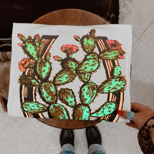 May include: White t-shirt featuring a vibrant cactus design. The illustration includes green cacti with brown accents and pink flowers, arranged in a horseshoe shape. The word "7" is also part of the design.