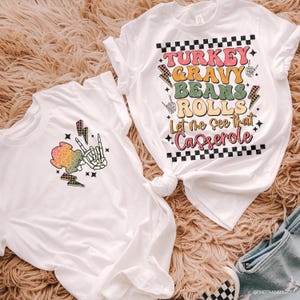 May include: Two white t-shirts with Thanksgiving graphics. One has a skeleton hand making a peace sign with a colourful cloud and lightning bolts. The other says "TURKEY GRAVY BEANS ROLLS Let me see that Casserole."