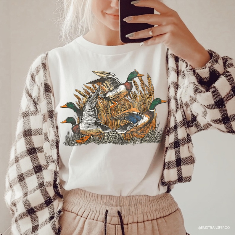 May include: White t-shirt featuring a detailed illustration of four ducks in a wheat field. The ducks have green heads, brown bodies, and blue wing accents. The shirt is worn with a black and white plaid jacket.