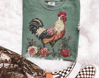 May include: A green t-shirt with a mosaic rooster design. The rooster is surrounded by pink roses and green leaves. The shirt is made of a soft, comfortable material.