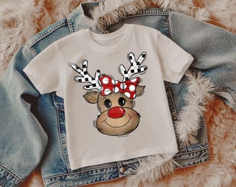 Puede incluir: Camiseta blanca de manga corta con un diseño de reno de dibujos animados. El reno tiene cuernos a cuadros blancos y negros, un lazo rojo con lunares y una nariz roja. La camiseta está sobre una chaqueta vaquera y una superficie suave de color marrón claro.