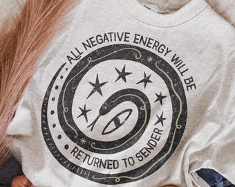 May include: A light gray crewneck sweatshirt featuring a black circular graphic with a coiled snake, stars, and the text "ALL NEGATIVE ENERGY WILL BE RETURNED TO SENDER" printed in a distressed, weathered font style across the center chest.