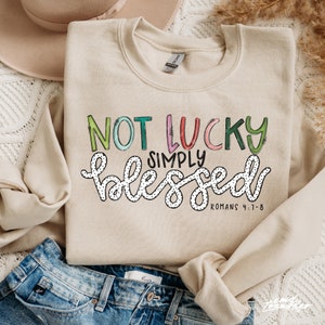 May include: Beige crewneck sweatshirt with the text "NOT LUCKY SIMPLY BLESSED" in colorful, whimsical lettering. The word "blessed" is in a cursive font with black polka dots. The text "ROMANS 4:1-8" is at the bottom.