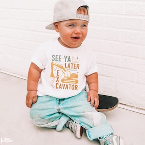 May include: A toddler wearing a white t-shirt with the text "See Ya Later Excavator" and a yellow excavator graphic. The toddler is wearing a grey baseball cap and blue jeans and is sitting on a skateboard.