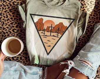 May include: A light green t-shirt with a graphic of a desert scene inside a black triangle. The design features brown rock formations, three crosses, and a setting sun. The shirt is on a leopard print surface.