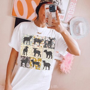 May include: White t-shirt with a patchwork design featuring black silhouettes of lambs and floral patterns. The design is centered on the shirt.