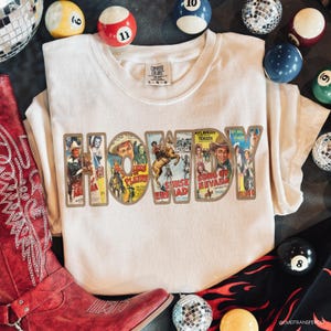 May include: A cream-colored t-shirt featuring the word HOWDY in large block letters. Each letter is filled with vintage Western movie poster imagery, including scenes of cowboys and horses. The shirt has a Comfort Colors label at the neck.