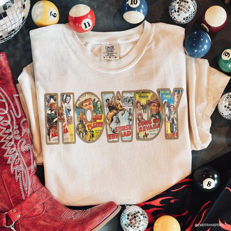 May include: A cream-colored t-shirt featuring the word HOWDY in large block letters. Each letter is filled with vintage Western movie poster imagery, including scenes of cowboys and horses. The shirt has a Comfort Colors label at the neck.