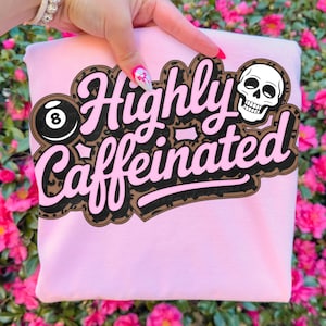 May include: A light pink sweatshirt with the words "Highly Caffeinated" in a black, brown, and pink design. The design includes an eight ball and a skull.