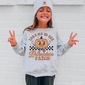May include: A light gray sweatshirt with the text "Take Me To The Pumpkin Patch" and a cartoon pumpkin graphic. The pumpkin has a smiling face and is wearing shoes. The sweatshirt is paired with a white beanie.