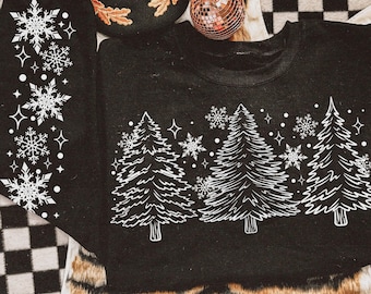 May include: A black long-sleeve sweatshirt featuring a white graphic print of three pine trees across the chest, surrounded by snowflakes and star shapes. The left sleeve also displays a vertical pattern of white snowflakes and star accents.