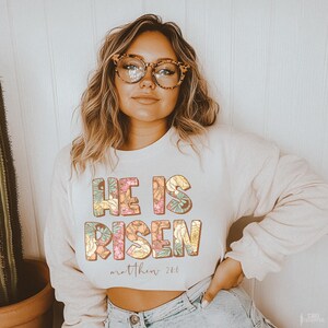 May include: A white cropped sweatshirt with the text "He is Risen" in colorful floral letters. The text "Matthew 28:6" is printed below the floral text.