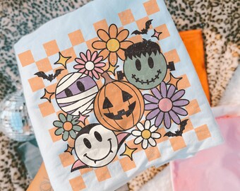 May include: A light blue t-shirt featuring a graphic print of a checkered orange pattern overlaid with a mummy, a jack-o'-lantern, a green Frankenstein monster, a vampire face, various colourful flowers, small bats, and four-pointed stars.