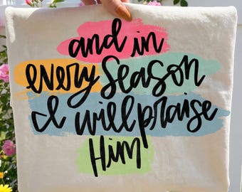 May include: Cream-colored canvas tote bag with the text "and in EVERY SEASON I will praise Him" in black script. The text is layered over colorful brushstrokes of pink, yellow, blue, and green.