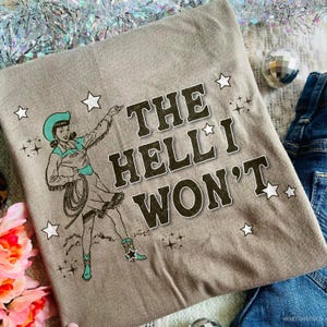 Puede incluir: Camiseta color topo con una ilustraci&oacute;n vintage de una vaquera con lazo, sombrero y botas turquesas. El texto "THE HELL I WON'T" est&aacute; impreso en una fuente desgastada, rodeado de estrellas. Un dise&ntilde;o divertido e informal.