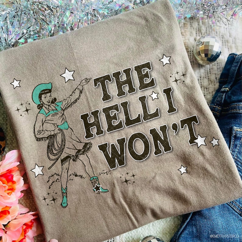 Puede incluir: Camiseta color topo con una ilustraci&oacute;n vintage de una vaquera con lazo, sombrero y botas turquesas. El texto "THE HELL I WON'T" est&aacute; impreso en una fuente desgastada, rodeado de estrellas. Un dise&ntilde;o divertido e informal.