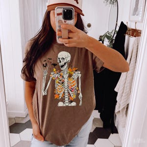 May include: A brown t-shirt with a skeleton graphic. The skeleton is surrounded by fall leaves and has a peace sign hand gesture. The shirt has a distressed look.