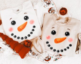 May include: Two long-sleeve crewneck sweatshirts, one white and one beige, each featuring a printed snowman face with black dot eyes, a textured orange carrot nose, and pink circular cheeks. The garments are arranged with holiday decor.