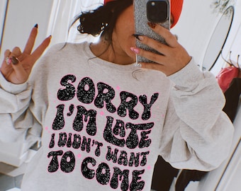 May include: A gray sweatshirt with the text "Sorry I'm Late I Didn't Want To Come" in black glitter letters. The letters are in a retro font style.