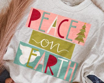 May include: A light grey crewneck sweatshirt featuring a graphic print with the text PEACE ON EARTH. The design includes a stylised green Christmas tree, a white dove silhouette, and colourful block lettering in shades of red, teal, and white.