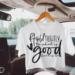 May include: White crop top with the text "Hold Tightly to what is good" in black script. The shirt has a fringe detail at the bottom and is hanging on a wooden hanger.