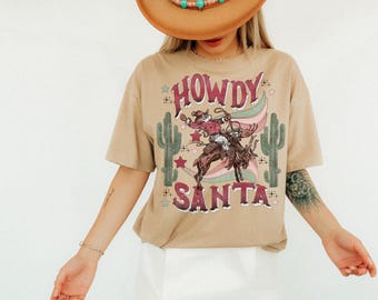 Può includere: T-shirt color cachi con grafica di Babbo Natale su un cavallo da rodeo, con la scritta "HOWDY SANTA" sopra e "SANTA" sotto. L'outfit è completato da un cappello da cowboy di paglia con perline turchesi e una gonna bianca.