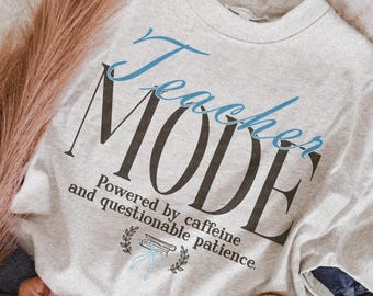 May include: A light gray crewneck t-shirt featuring the blue script text Teacher and the large black serif text MODE. Below, the black text reads Powered by caffeine and questionable patience, accompanied by a small blue book graphic.