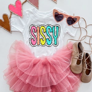 May include: A white t-shirt with the word "SISSY" printed in colorful letters with black polka dots. The shirt is paired with a pink tulle tutu and brown leather shoes.