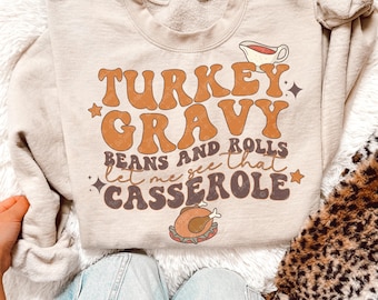 May include: Beige sweatshirt with the text "TURKEY GRAVY BEANS AND ROLLS Let me see that CASSEROLE" in brown and orange lettering. The design includes a gravy boat, stars, and a roasted turkey illustration. The sweatshirt is a cozy, casual style.