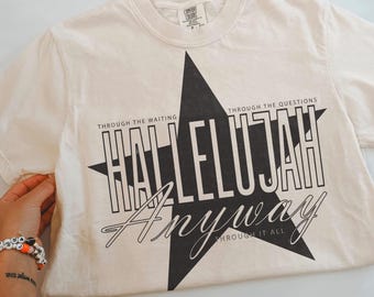 May include: An off-white short-sleeve t-shirt featuring a large black star graphic. The text reads "THROUGH THE WAITING," "HALLELUJAH," "THROUGH THE QUESTIONS," and "Anyway THROUGH IT ALL" in a mix of bold block and cursive lettering.