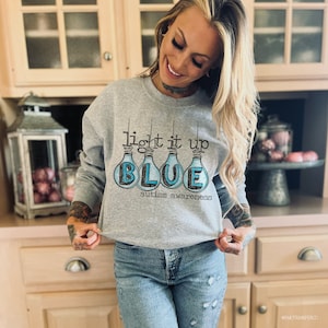 May include: A gray sweatshirt with the text "light it up BLUE autism awareness" and a graphic of light bulbs with the word "BLUE" inside.