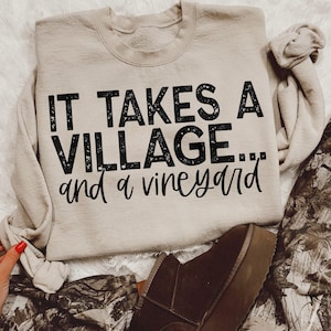 May include: A beige sweatshirt with the text "It takes a village... and a vineyard" printed in black.