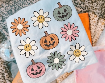 May include: A light blue t-shirt featuring a repeating graphic print of three jack-o'-lantern pumpkins in green, orange, and pink, interspersed with six daisy flowers in white, pink, and green, each with a yellow smiley face centre.